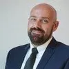 A person with a shaved head and facial hair, wearing a dark blue blazer, white shirt, and dark textured tie.