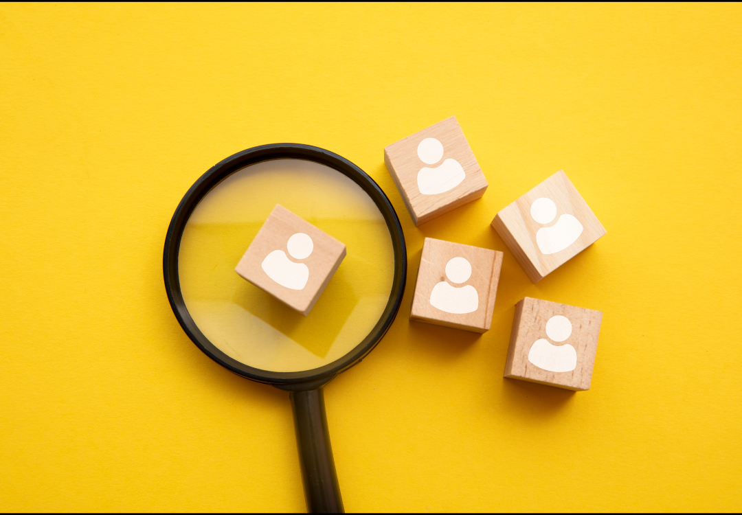A magnifying glass focuses on one of five wooden blocks, each featuring a white human-shaped icon on a yellow background.