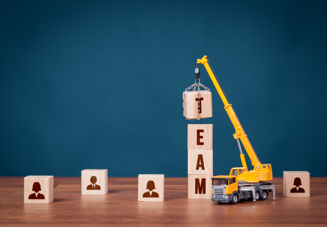 A toy crane lifts a wooden block marked with 'T' onto a stack spelling 'TEAM' surrounded by blocks with person icons.