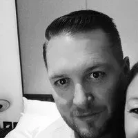 A close-up, black-and-white portrait of a man looking toward the camera, partially cropped next to another person.