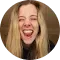 A person with long blonde hair laughing with their mouth wide open against a brown background.