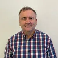 A person with short hair wearing a navy, white, and red plaid button-down shirt against a plain, light-colored background.