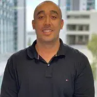 A person in a dark polo shirt smiles, centered in front of a blurred background of a modern office building.