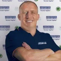A smiling professional with arms crossed in front of a white wall patterned with a construction recruitment logo.