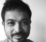 A black and white portrait of a person with short hair and a beard, looking directly at the camera with a soft smile.