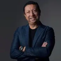 A person wearing a dark blue blazer and black shirt, smiling with arms crossed, set against a dark gray background.