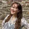 A smiling person with glasses and long, wavy hair wears a patterned shirt in front of a textured stone wall.