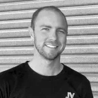 A smiling person wearing a dark t-shirt, posed in front of a light-colored, horizontally-ribbed metal wall.
