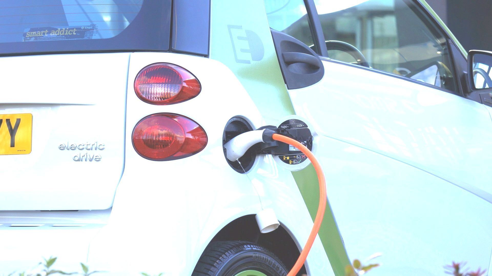 A white electric smart car parked outdoors, charging via an orange cable plugged into its side.