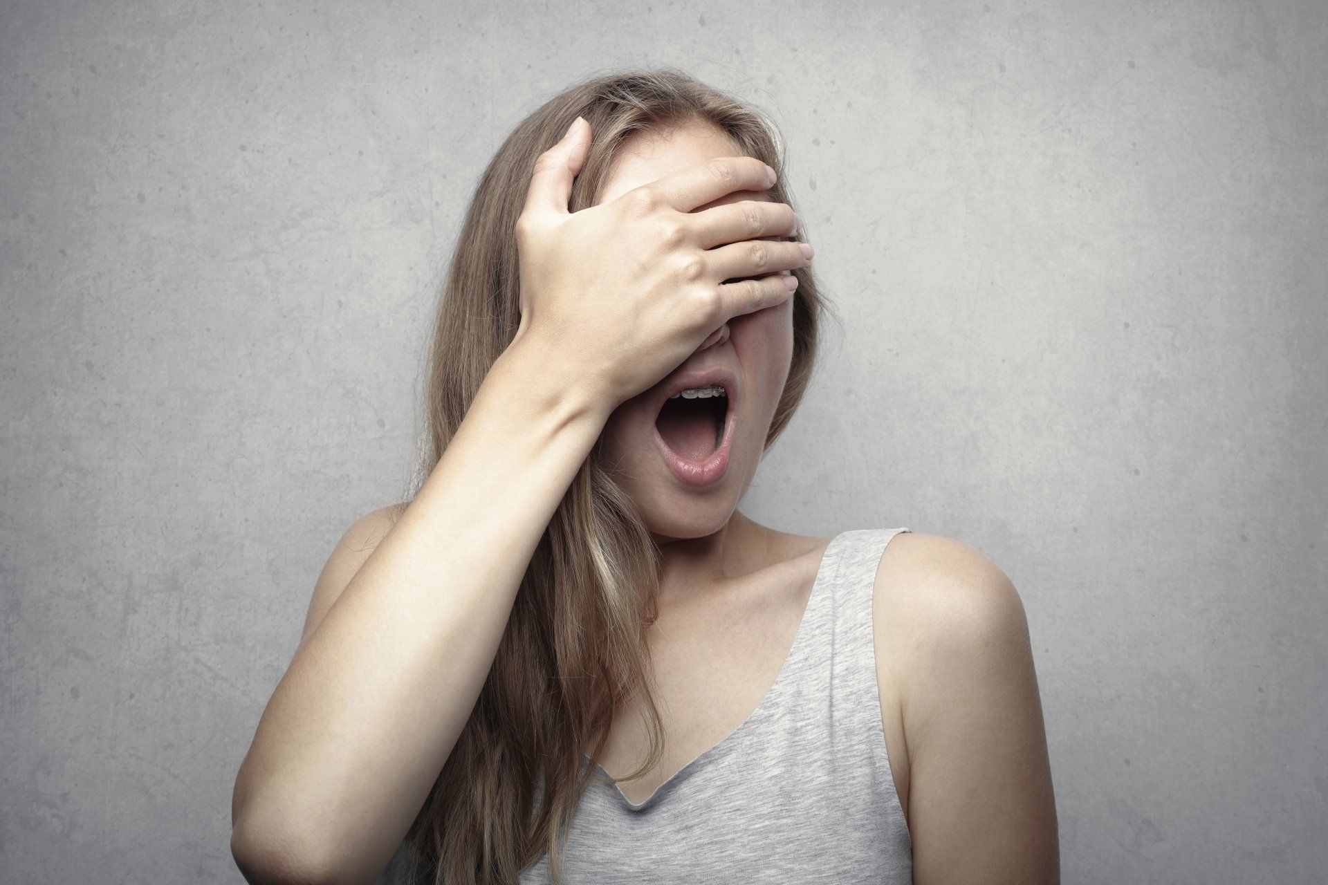 A person with long hair wearing a grey tank top covers their eyes with one hand while their mouth is open in a yawn.
