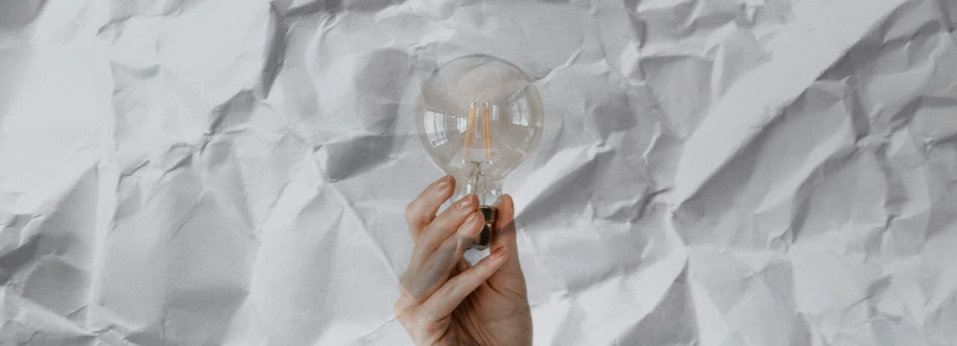 A hand holds a clear, glowing lightbulb against a background of crumpled white paper.