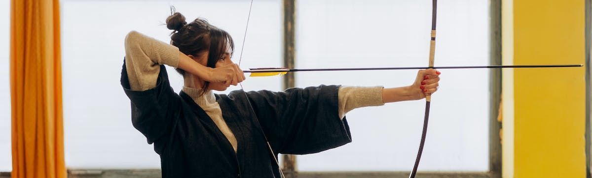 An archer in traditional dark clothing takes aim with a bow and arrow in a bright room with yellow curtains.