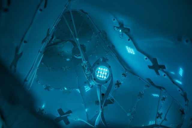A futuristic, blue-lit tunnel or conduit with geometric cables and circular lights illuminating the space.