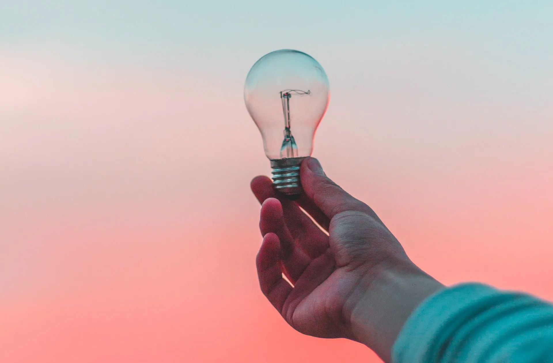 A person holds an unlit glass lightbulb against a soft, gradient pink and blue sunset background.