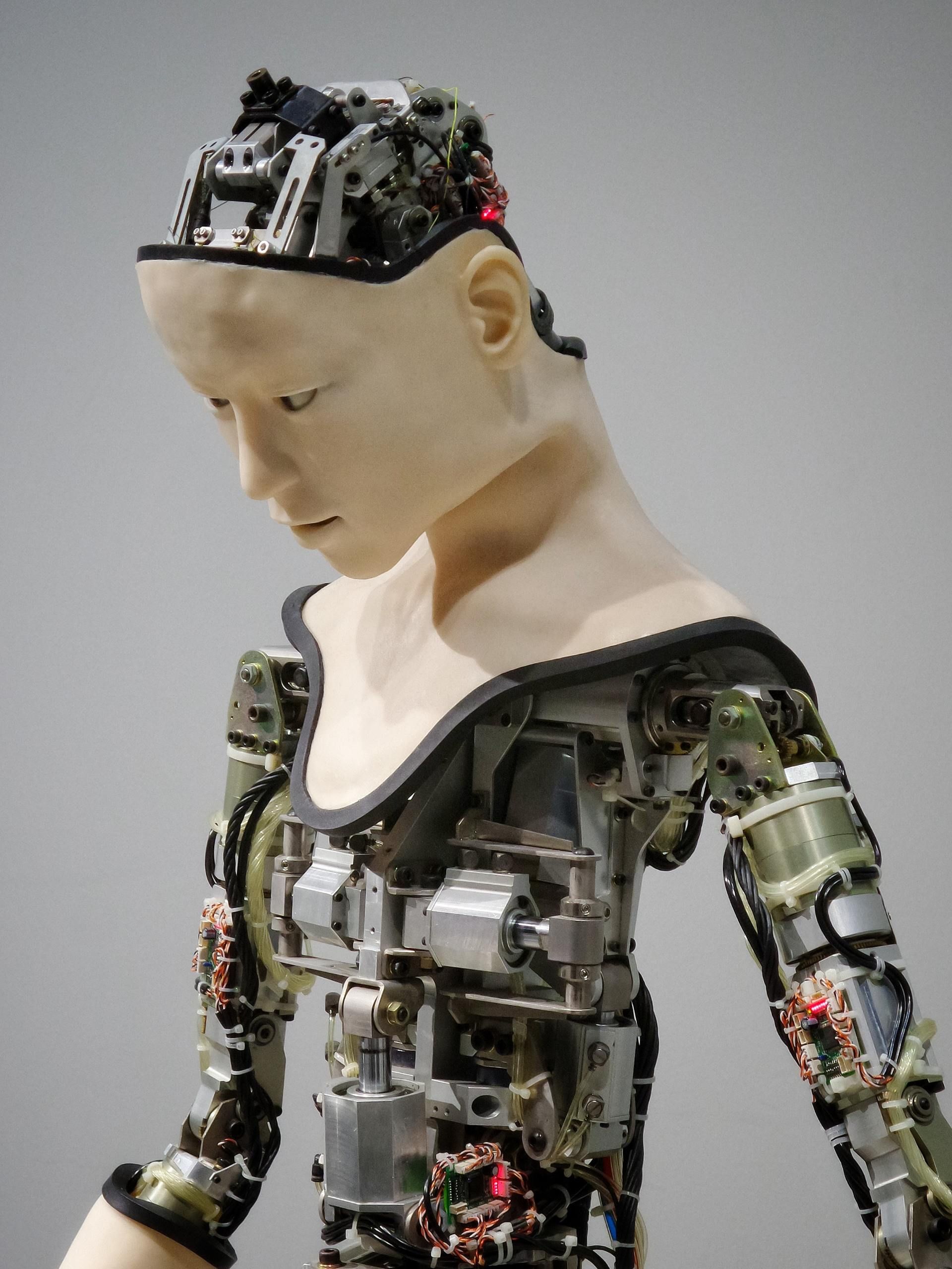 A robotic figure with a human-like face and exposed mechanical chest and arm components against a plain gray background.
