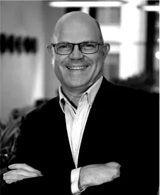 A smiling professional wearing glasses and a suit jacket, posing with arms crossed in an office setting.