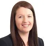 A professional headshot of a smiling person with shoulder-length brown hair, wearing a dark blazer against a white background.