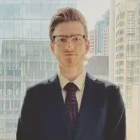 A person with short, light-brown hair and glasses wears a dark suit and tie, standing in front of a window with city views.