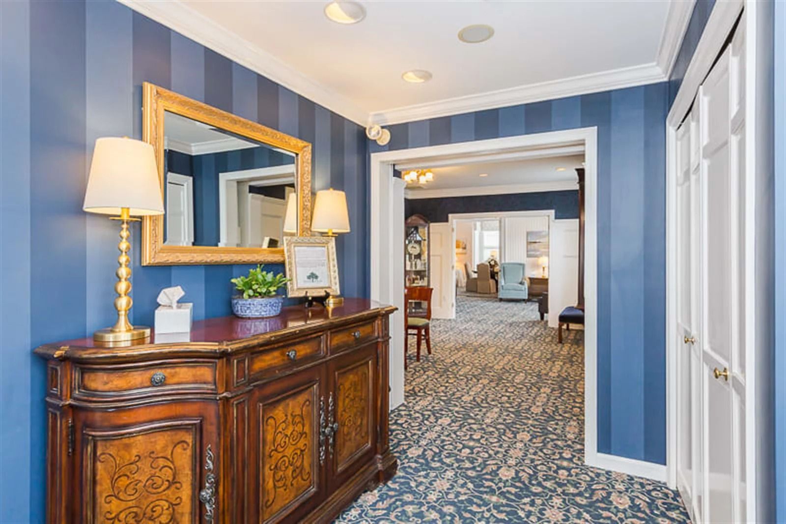 Blue-striped hallway with a wooden console table, gold lamp, mirror, and patterned carpet leading to a room