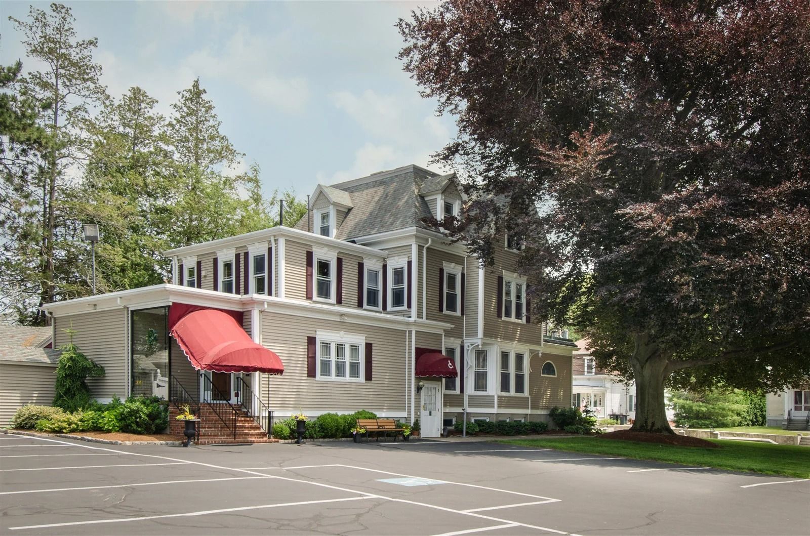 Two-story tan building with red awnings beside a parking lot and large trees