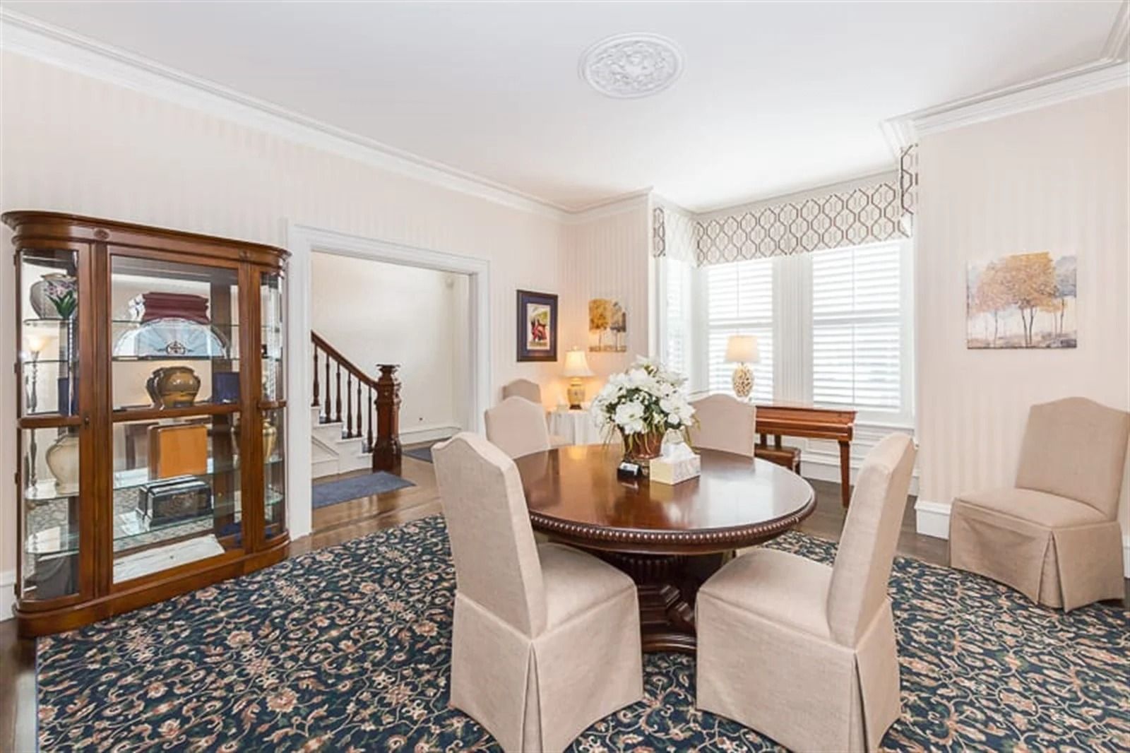 Bright dining room with glass cabinet, round table, beige chairs, and patterned carpet by a window