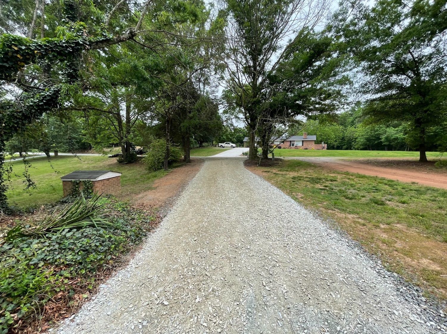 Gravel driveway leads toward a brick house, bordered by trees and green grass on either side.