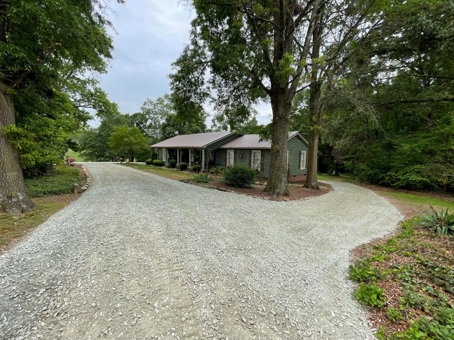 Gravel driveway splitting to approach a small, green house with a brown roof. Trees surround the house.