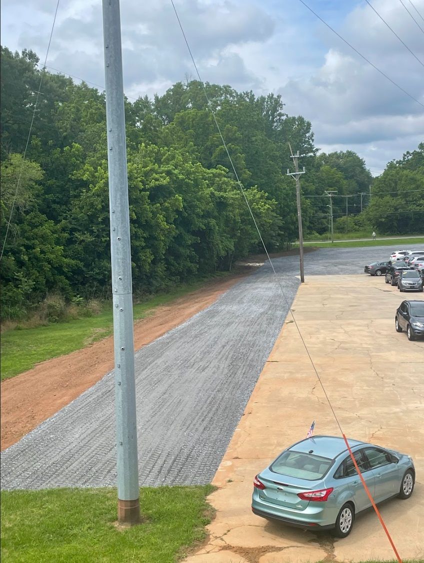 Gravel road next to a concrete parking area with a teal car. Power poles and trees in the background.