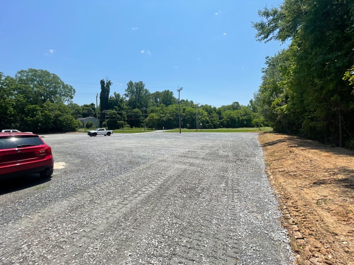 Red car parked on gravel lot under blue sky, surrounded by trees.