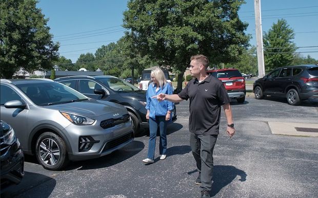 Two people walking beside parked cars in a sunny parking lot, one holding papers