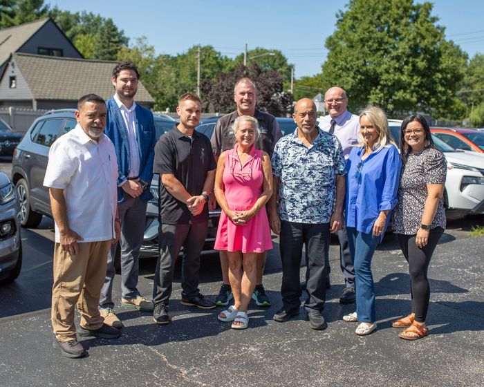 Group of nine people standing outdoors in a parking lot in front of houses, smiling at the camera.