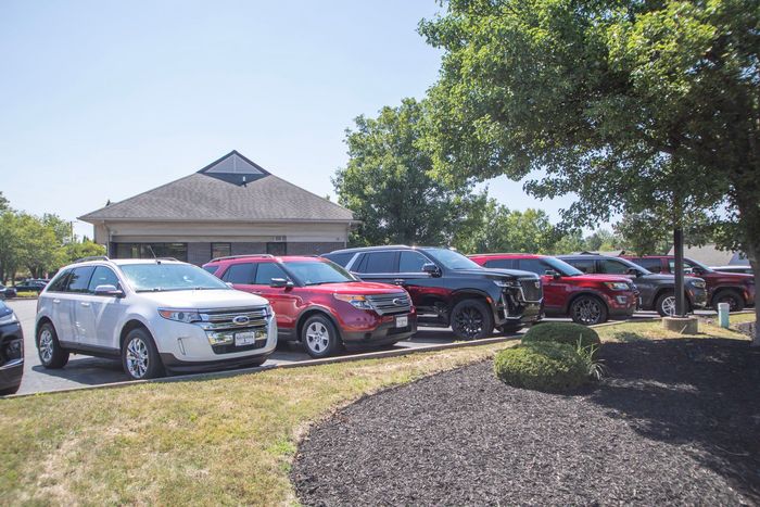 Row of parked cars beside a building on a sunny day, with trees and grass in the foreground