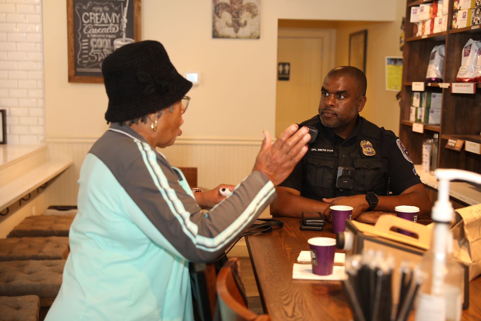 Person in turquoise jacket talks to a seated police officer at a café counter.