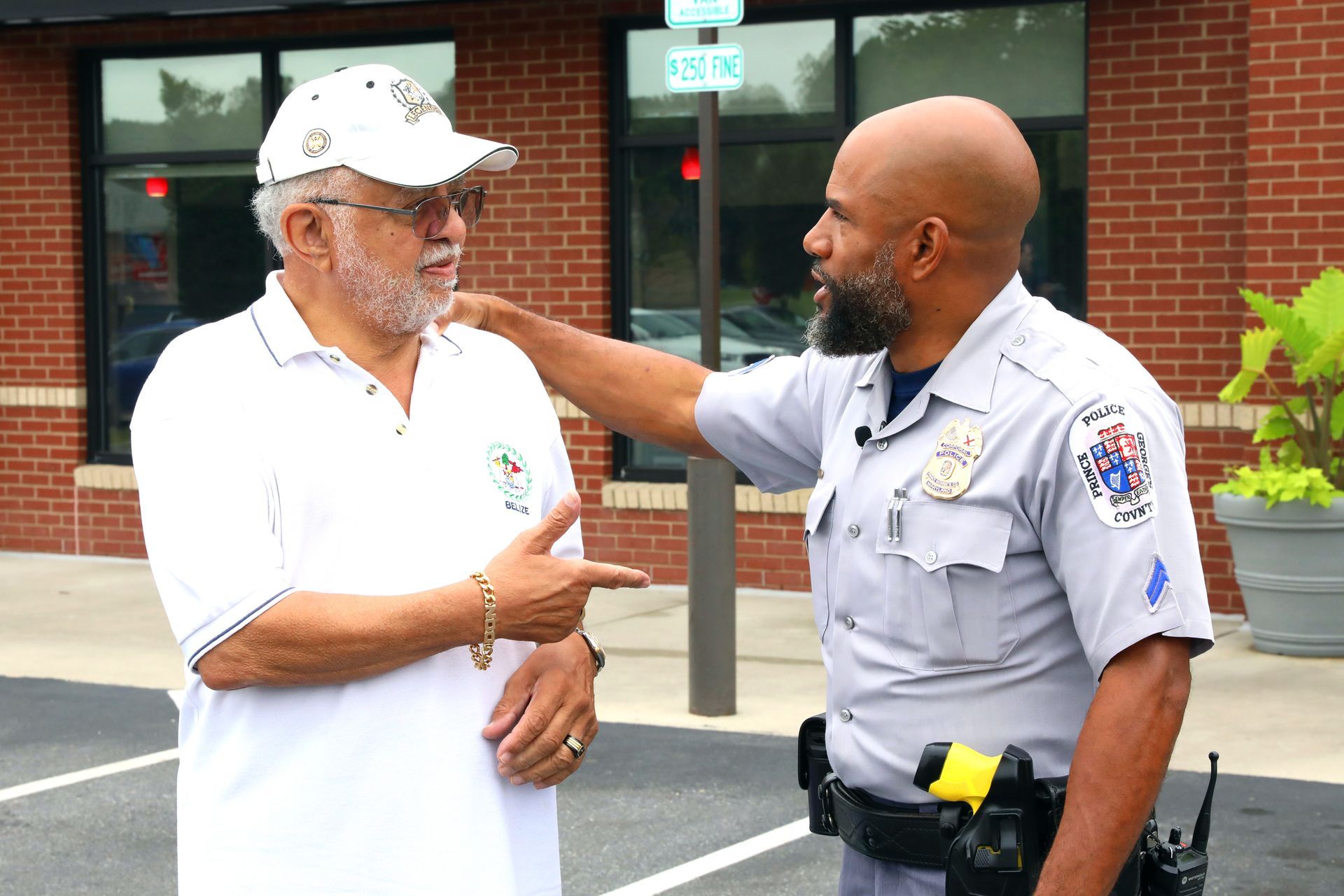 Two men speaking outside a building, one in a white polo and one in a gray uniform, with a red brick wall behind them