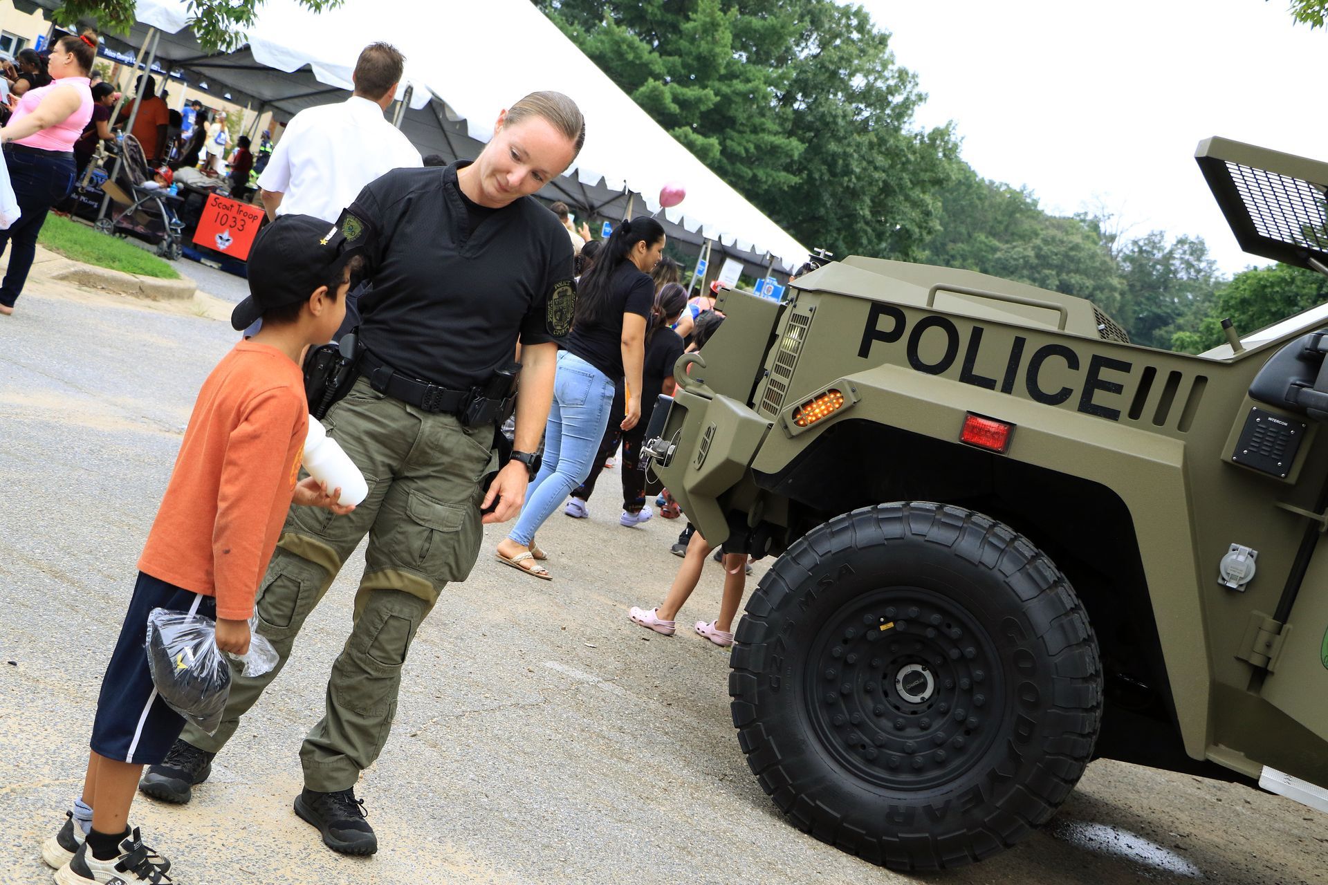 Police vehicle at an outdoor event as a man walks beside a child near a crowd and tents