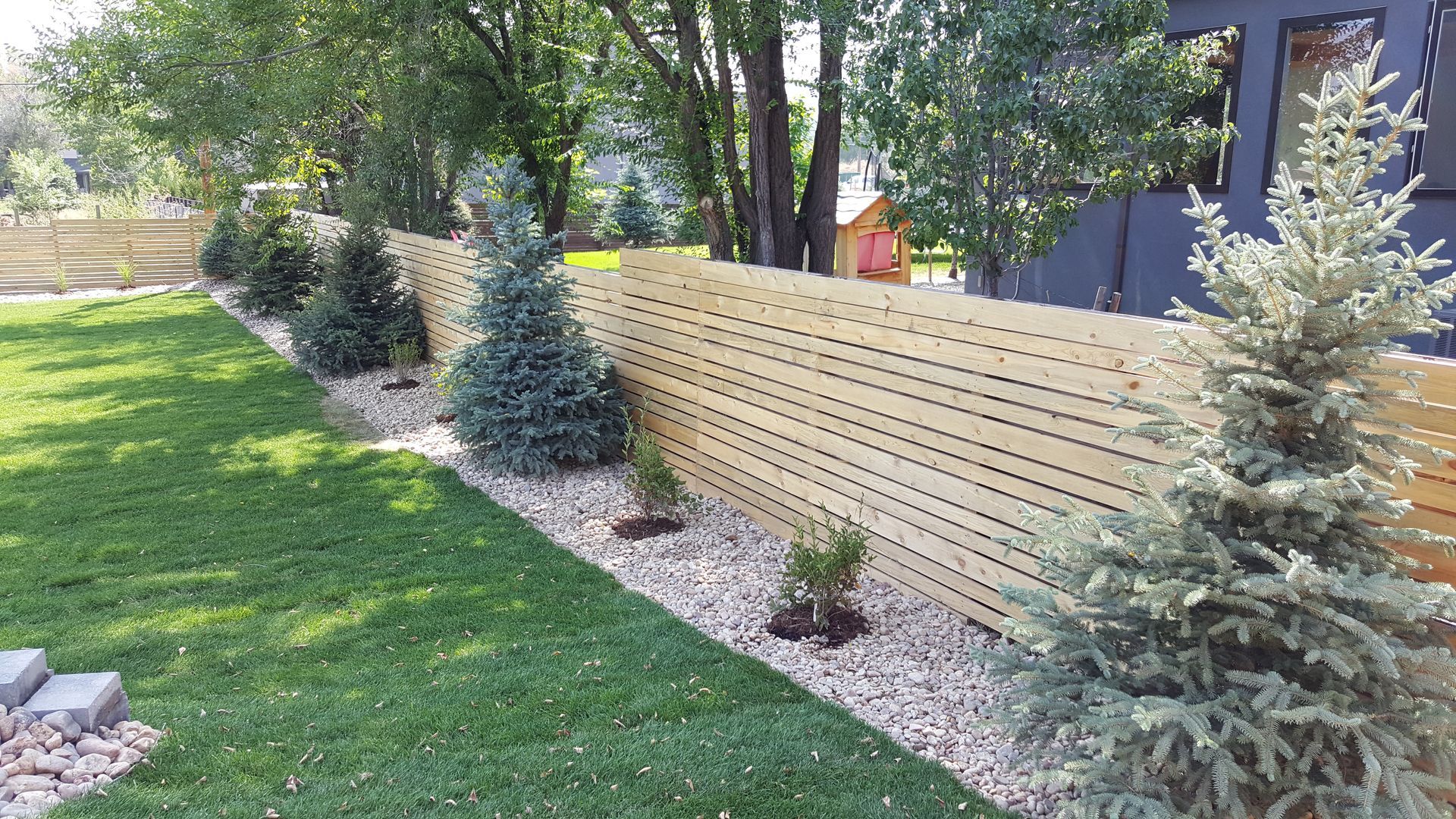 A wooden fence built by Greater Western Fence surrounds a lush green yard with trees and bushes.