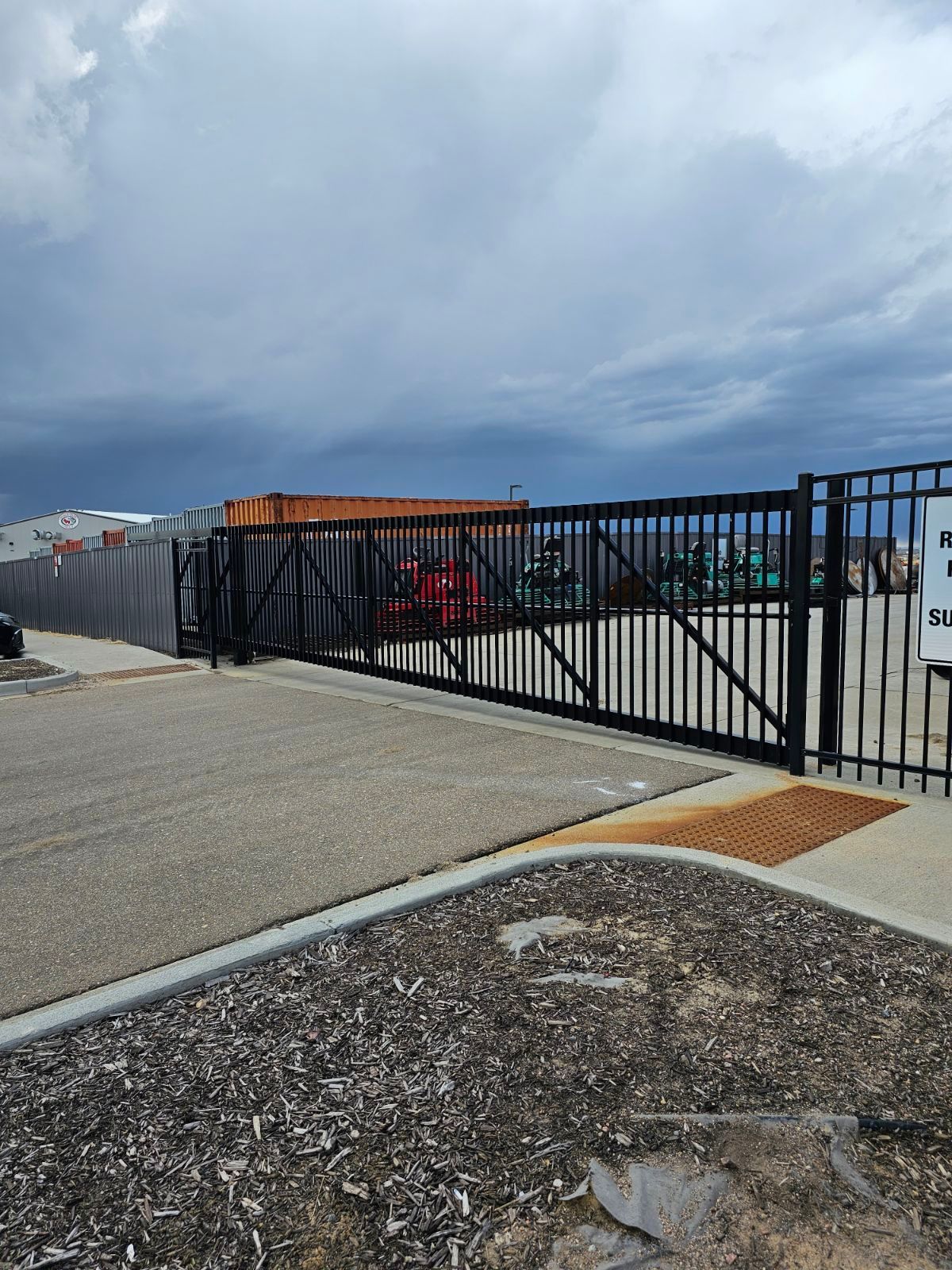 A black fence built by Greater Western Fence surrounds a parking lot with a cloudy sky in the background.