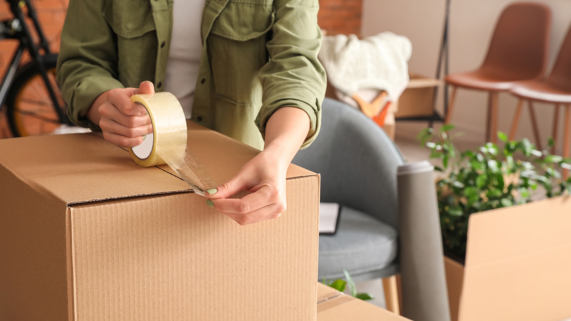 A person sealing a moving box with packing tape, preparing for a move in a well-organized living space with plants and furniture in the background.