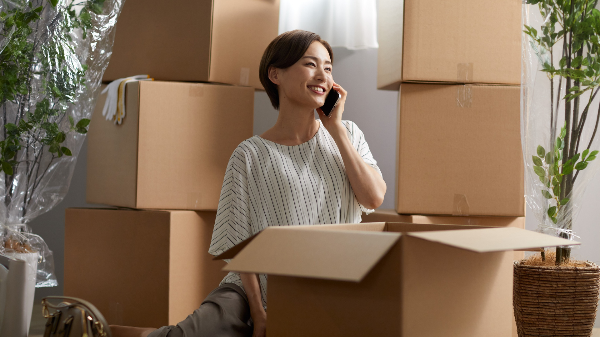 A smiling person talks on a smartphone, sitting surrounded by cardboard moving boxes in a bright, indoor setting.
