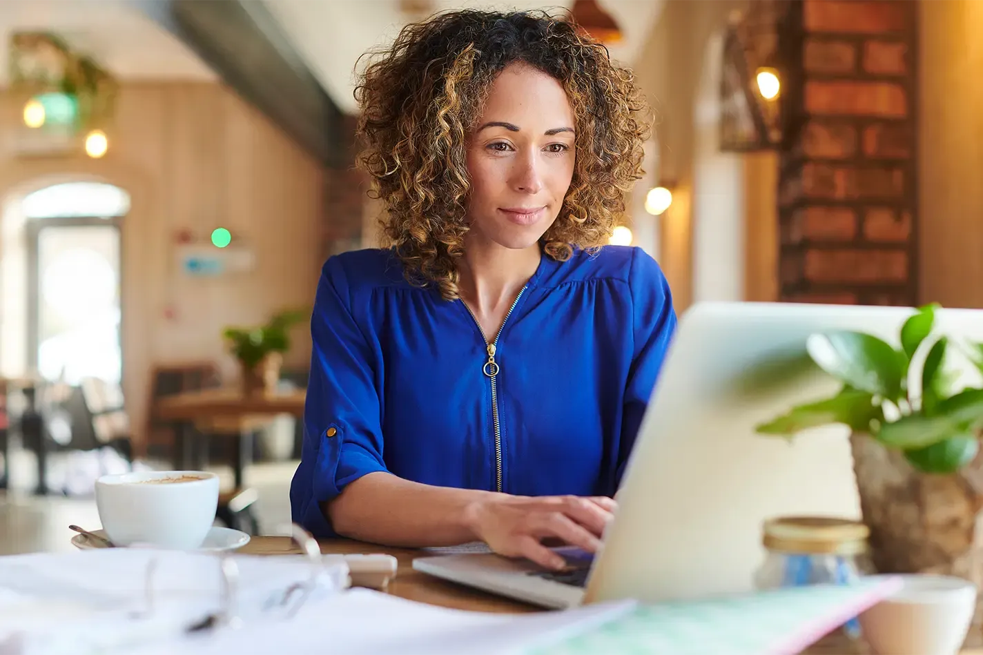 Woman at a cafe, typing on a laptop, with a cup of coffee and papers on the table.
