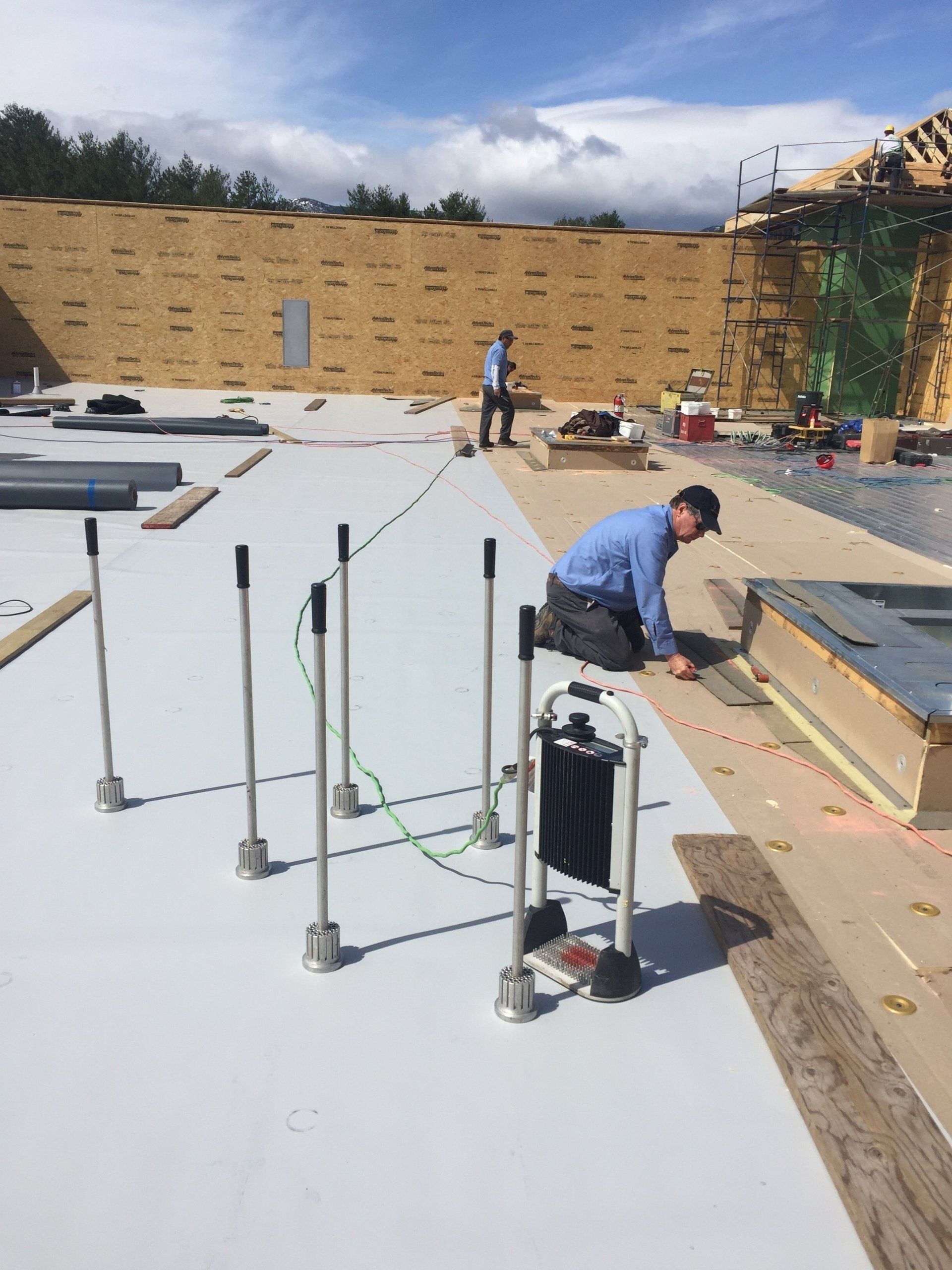 Roofer working on a flat rooftop with white panels and tools, near a tall tan wall and blue sky