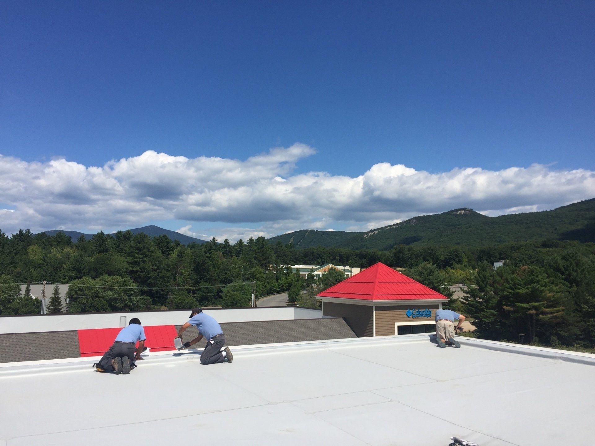 Three people work on installing a white flat roof on a building against a background of mountains and a blue sky.
