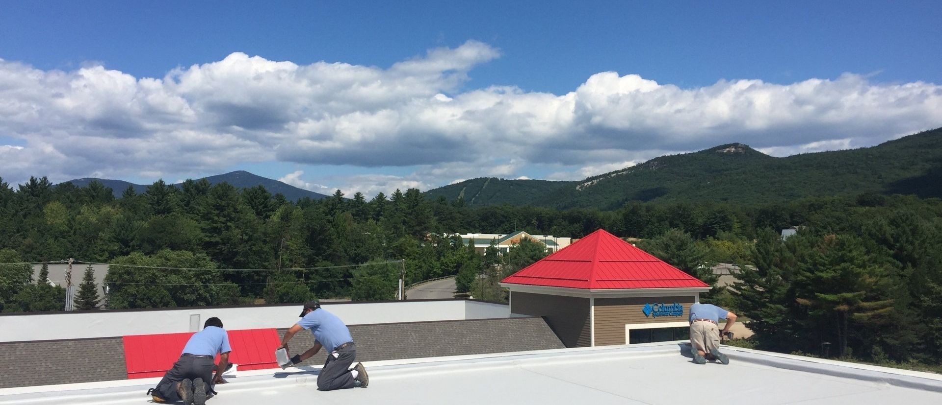 People kneel on a rooftop overlooking green mountains and a red-roofed building under a cloudy sky