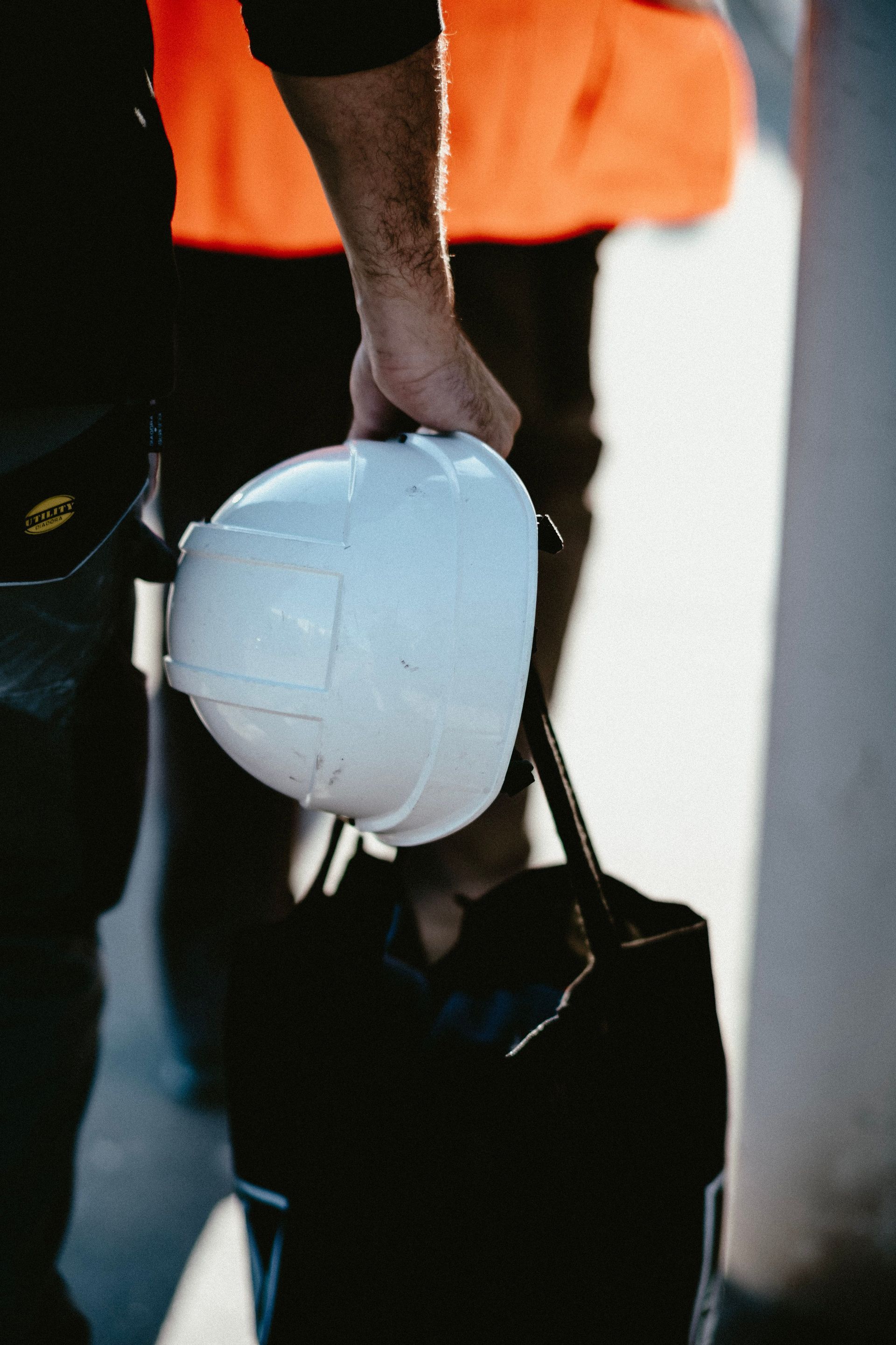Worker holding a white hard hat over a black bag, with an orange vest in the background