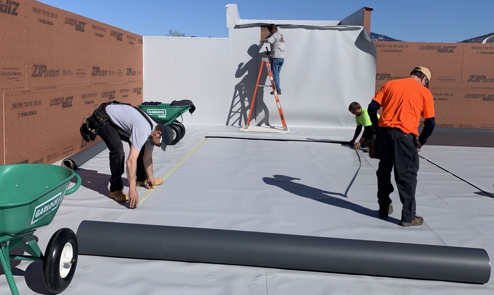 Workers installing a white rooftop membrane on a flat roof, with rolls of material and ladders nearby.