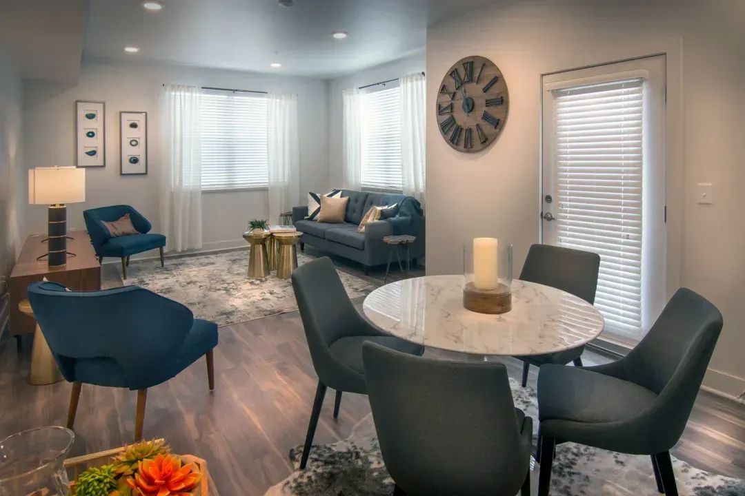 Bright modern living room with blue chairs, marble dining table, sofas, wall clock, and tall windows