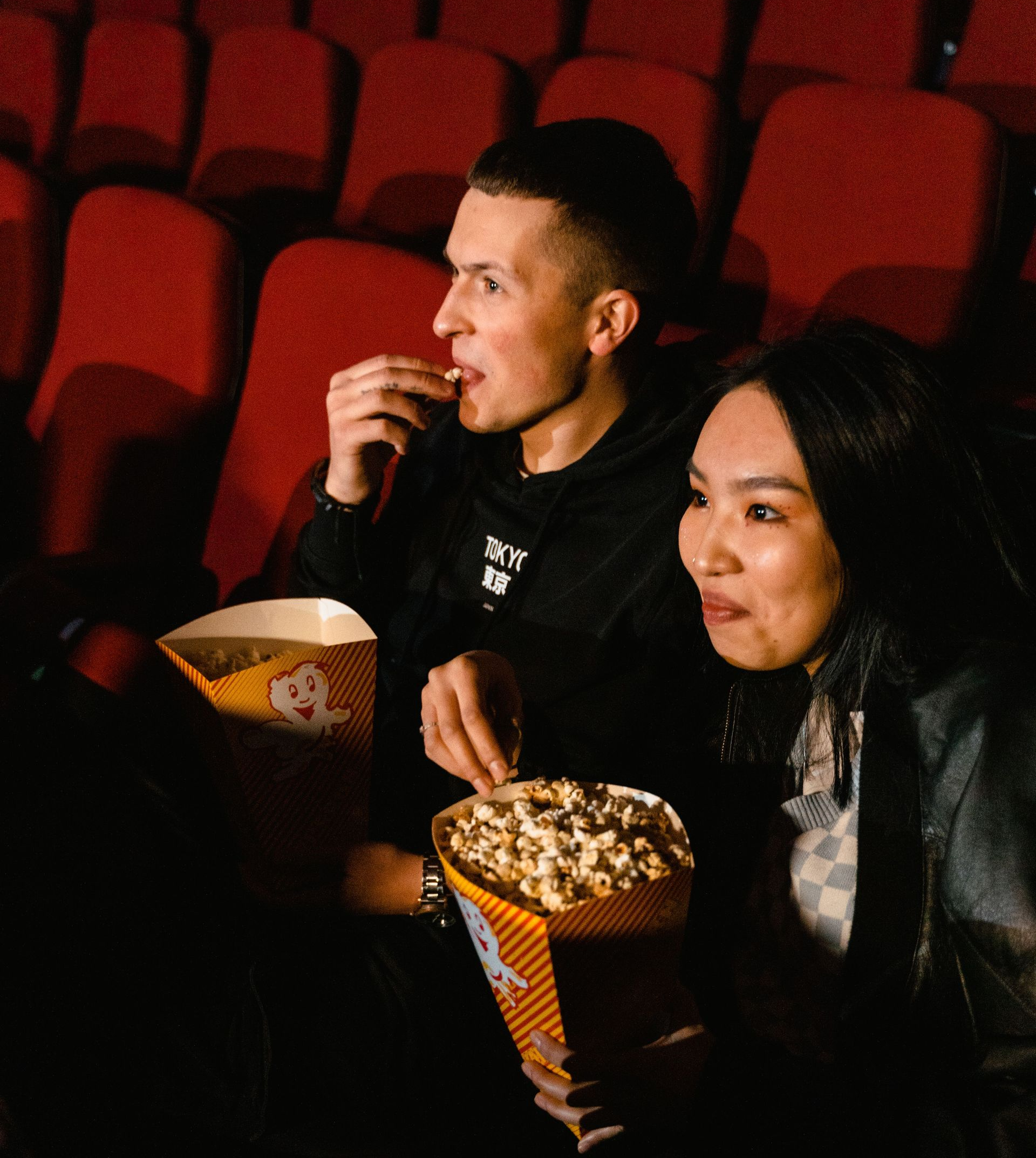 Two people seated in a dim theater, eating popcorn from red-and-white striped tubs