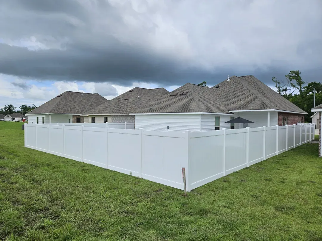 Single-story homes with gray roofs behind a white fenced yard on a grassy lot under cloudy skies