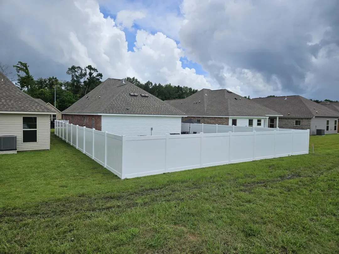 Backyard with white vinyl fence around a grassy yard beside single-story homes under a cloudy sky
