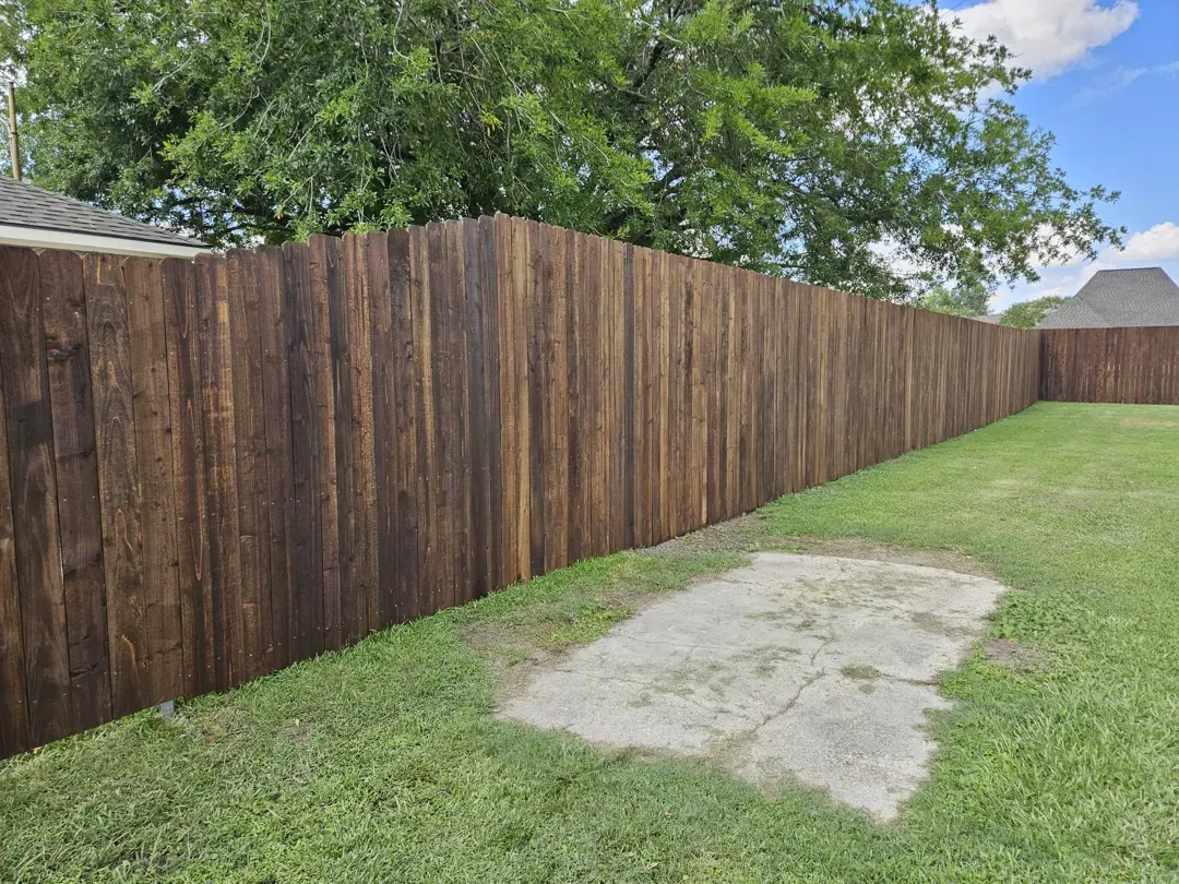 Long wooden privacy fence beside a grassy yard with a small concrete patch and trees overhead